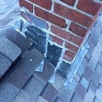 Close-up of a brick chimney on a shingled roof, showing metal step flashing sealed with caulk along the side.