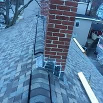 A high-angle view of a grey shingled roof with a brick chimney and metal flashing, overlooking a house and yard.