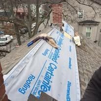 A roof under repair, featuring laid CertainTeed synthetic underlayment, a chimney, and residential buildings in the back.
