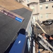 A roofer works on a sloped roof edge near a white building, with Landmark Pro shingle packaging visible on the deck.