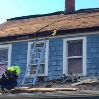 A worker in high-visibility gear kneels on a residential roof, repairing shingles near a ladder against a blue house.