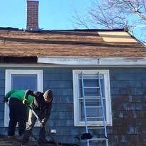 Two people work on a residential roof repair, with one person kneeling and another standing near a ladder.