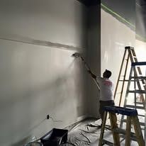 A person in a white shirt paints a large, smooth wall using a long-handled roller, standing near a tall metal ladder.