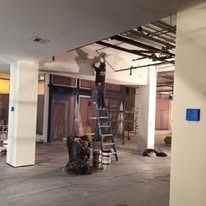 A worker on a ladder installs or repairs ceiling fixtures in a commercial space undergoing construction.