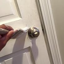 A hand uses a paintbrush to apply white paint to a wooden door around a metallic doorknob.