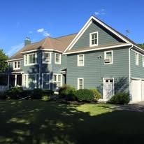 A two-story house with dark green siding, white trim, a brown roof, and a green lawn under a clear blue sky.