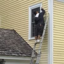 A person in a dark jacket and shorts stands on an extension ladder, working on a window frame on a yellow-sided house.