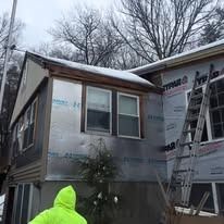 A person in a bright neon yellow coat stands outside a house undergoing construction, featuring silver-wrapped siding.