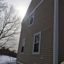 A side view of a tan, vinyl-sided house with three rectangular windows, against a bright, hazy sky and a snowy yard.