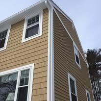 Tan vinyl-sided house exterior showing a corner with white trim, windows, and dark-colored siding shingles.