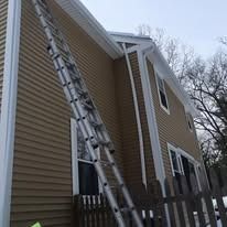 A long extension ladder leans against the side of a beige, two-story house with white trim and a wooden fence.