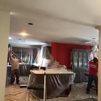 Two workers prepare a residential interior for painting, with furniture covered in plastic sheeting.
