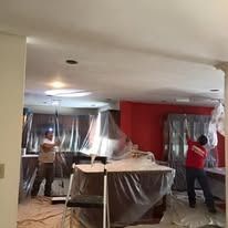 Two people in a kitchen covered in plastic sheeting for painting, working near a bright red accent wall.
