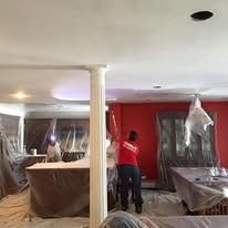A person in a red shirt paints a ceiling in a room with white columns, red walls, and plastic-covered furniture.