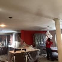 A person in a red shirt works on a ceiling in a home interior covered in protective plastic sheeting.