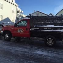 A red utility truck with a black dump bed parked on a snow-covered street in front of a white building.
