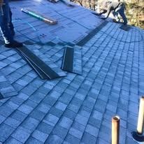 A worker installs gray asphalt shingles on a roof, with tools and materials visible on the paper underlayment.