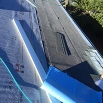 A close-up of a roof under construction, showing gray felt underlayment and a section of installed black roofing material.