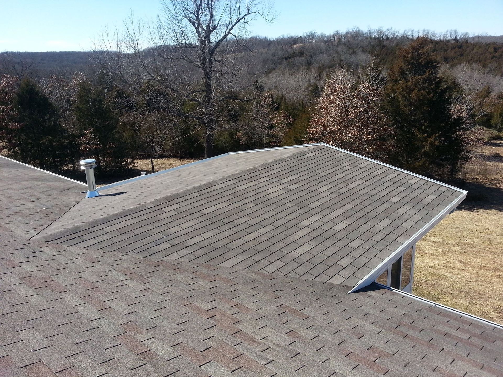 Brown shingled roof of a house with a scenic, tree-filled backdrop on a sunny day.