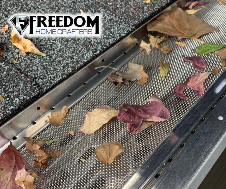 Gutter guard with leaves; close-up view. Silver metal mesh, red and brown leaves, gray rooftop.