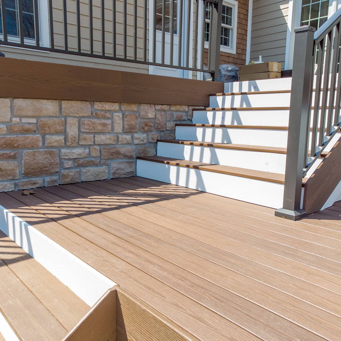 Wooden deck with stairs leading to a house, stone wall, and brown and white accents.