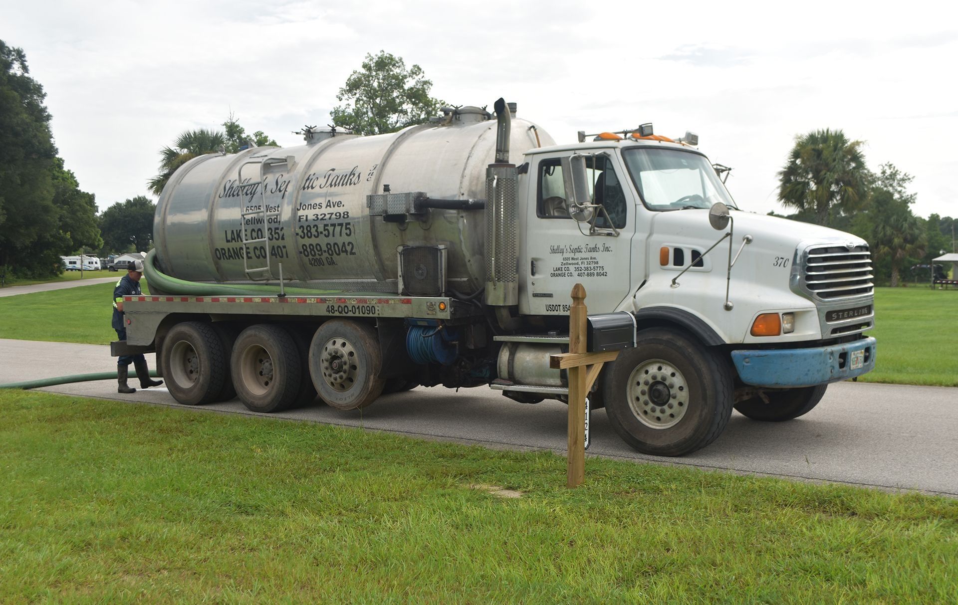 White tanker truck parked on a road beside a grassy area and mailbox under cloudy skies