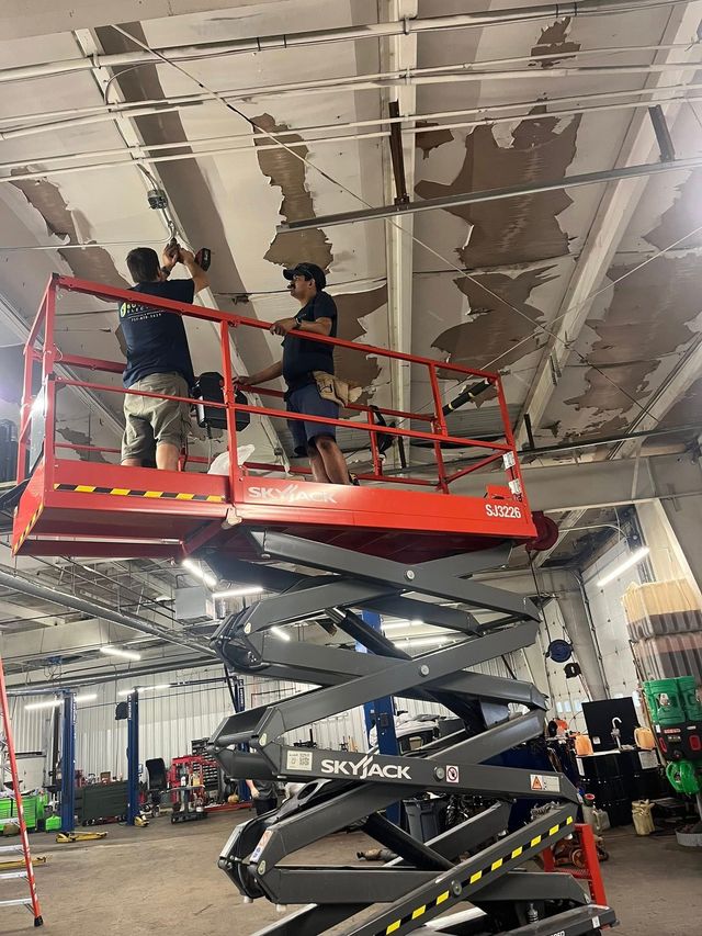 Two people on a red scissor lift, installing something on a ceiling.  Large room with exposed beams.