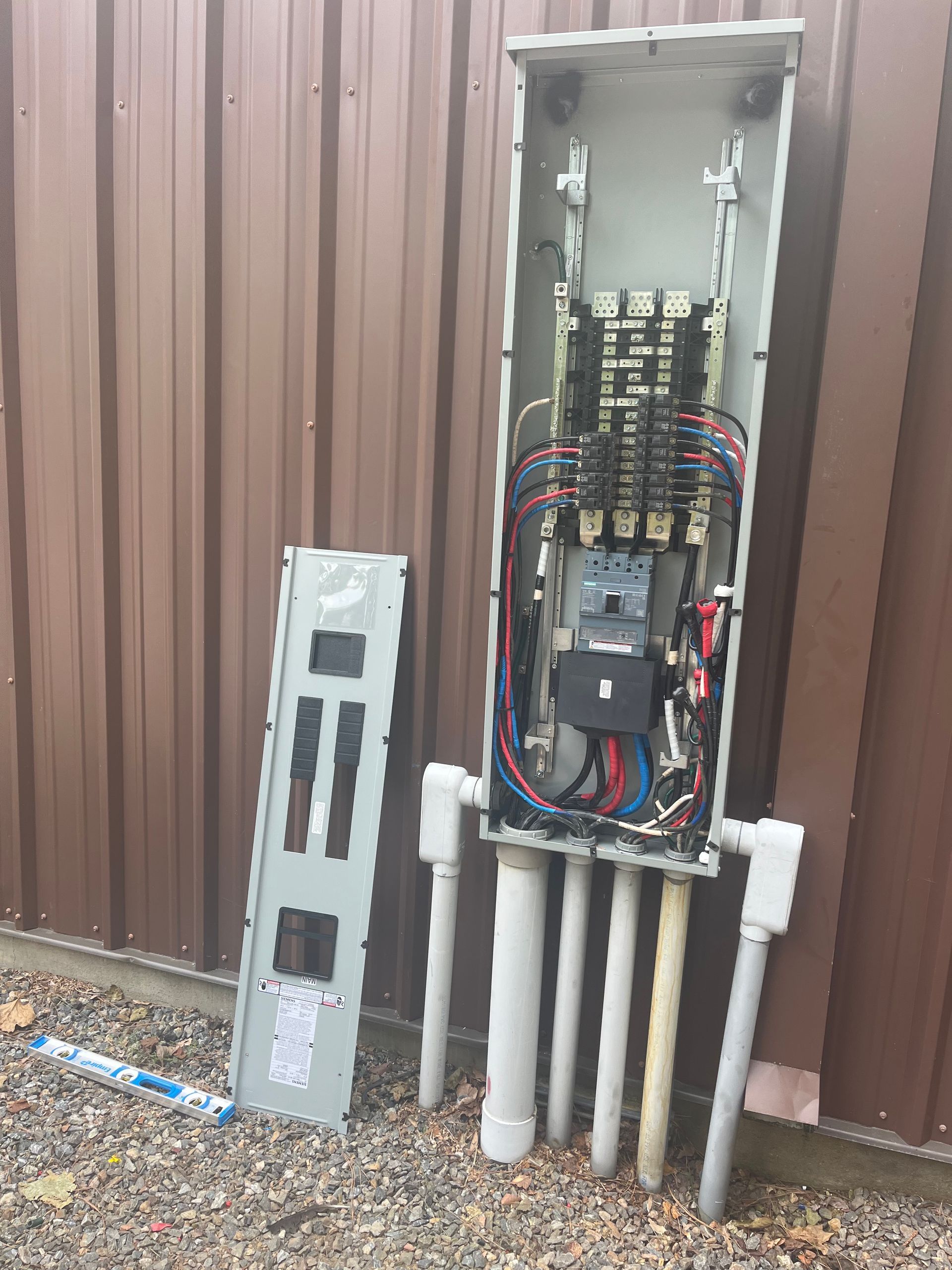 Open electrical panel with cover on ground, beside brown wall, conduit pipes below.