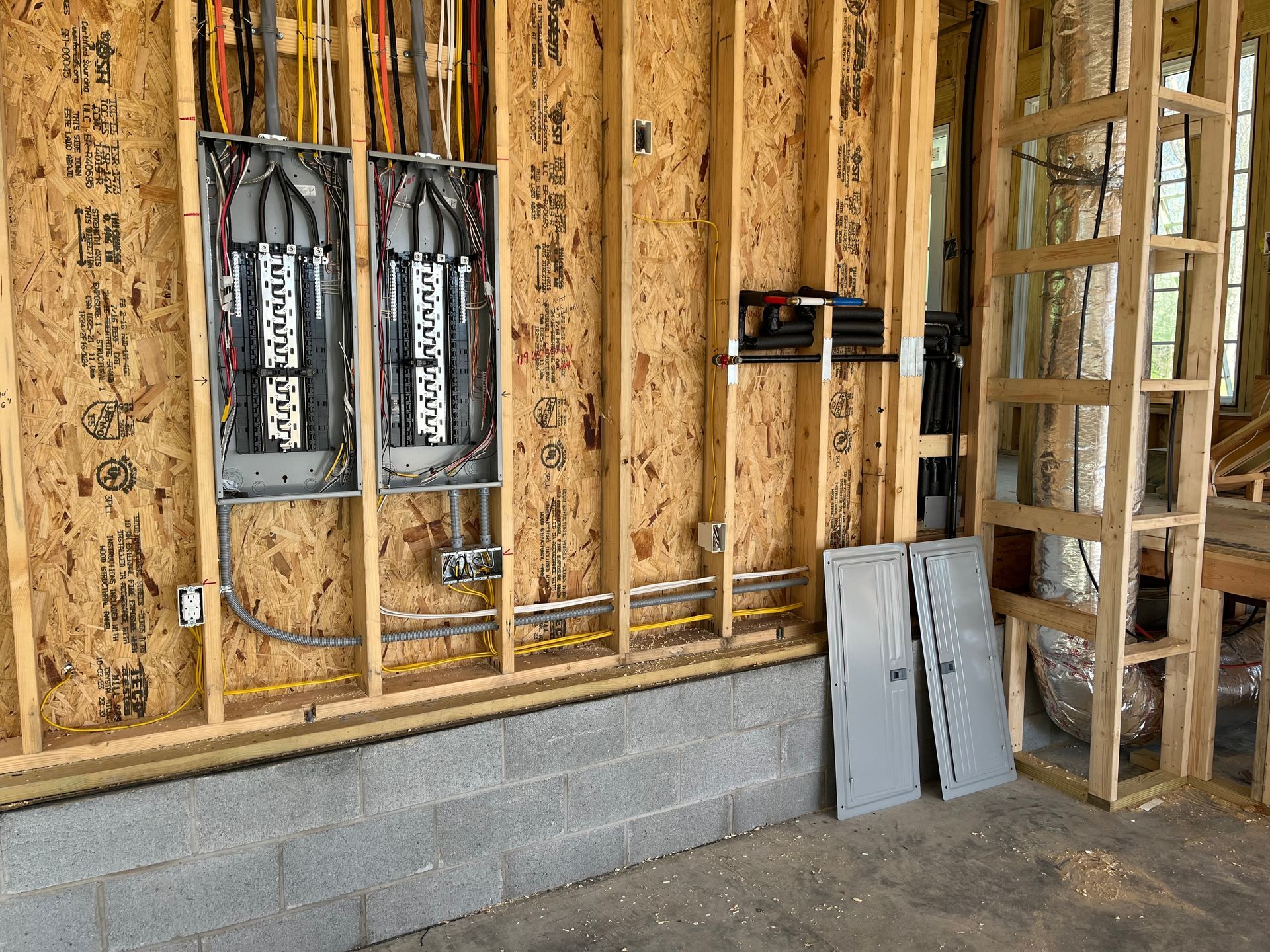 Electrical panel installation in a wood-framed wall. Gray panels lie below. Conduit and wiring are visible.
