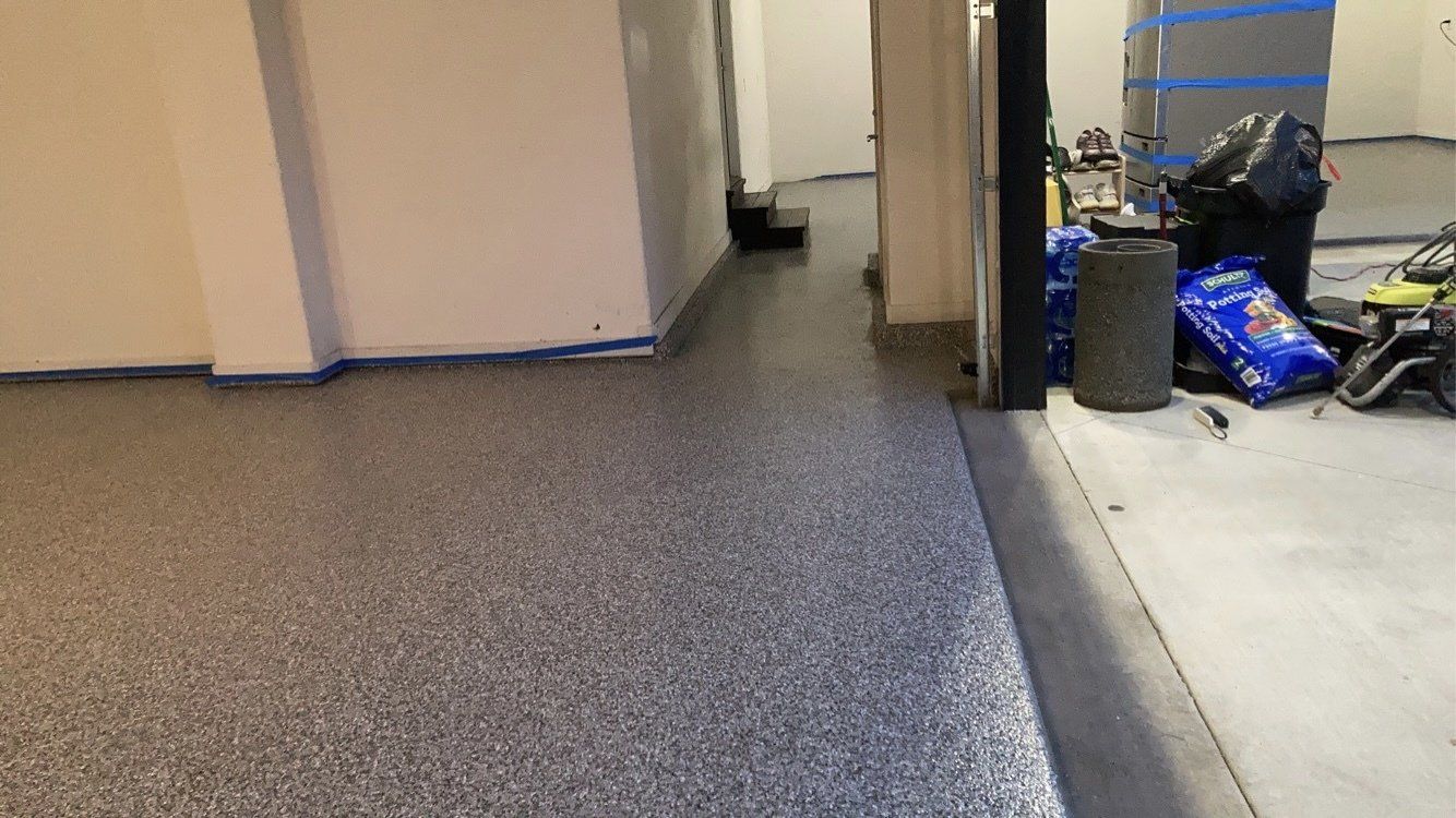 A concrete floor with gray and silver speckles. A doorway leads to a cluttered area with tools and a trash bag.