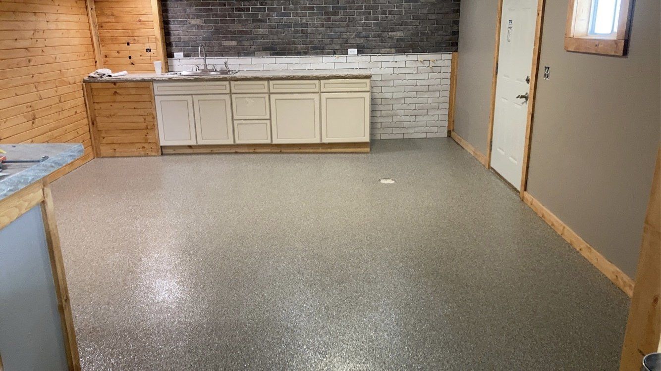 A finished basement with a gray speckled epoxy floor, white cabinets, and exposed brick wall. The room has a door and a small window.