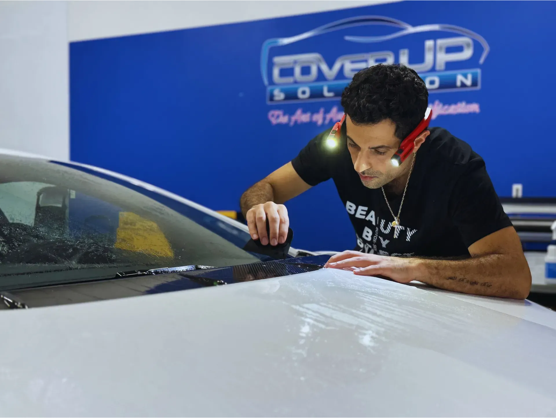 A technician with headlamps inspecting a vinyl wrap being applied to the hood of a white car in a shop.
