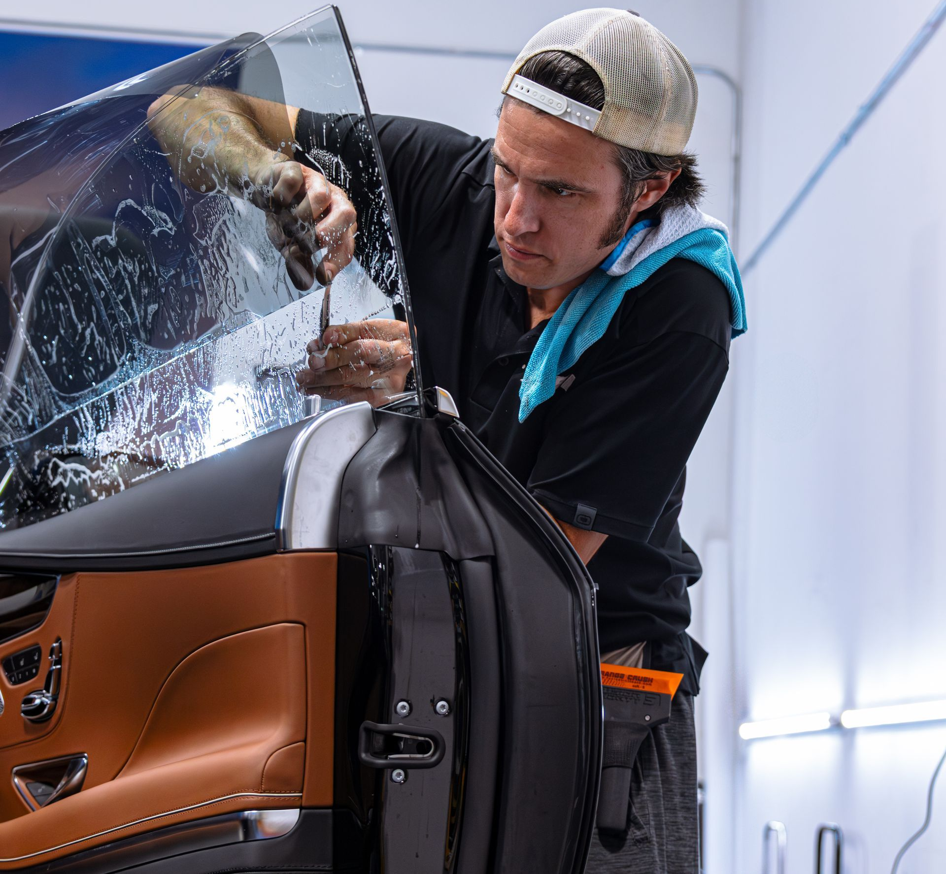 Person tinting a car window; using a tool, wiping sweat. Tan interior, black car door, light setting.