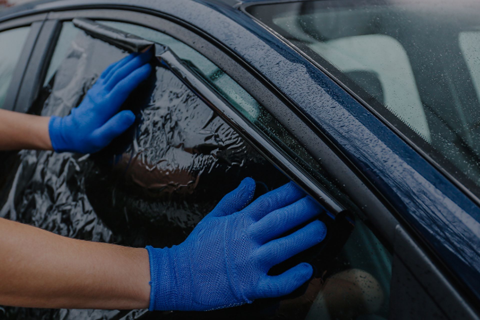Hands in blue gloves applying tinted film to a car window.