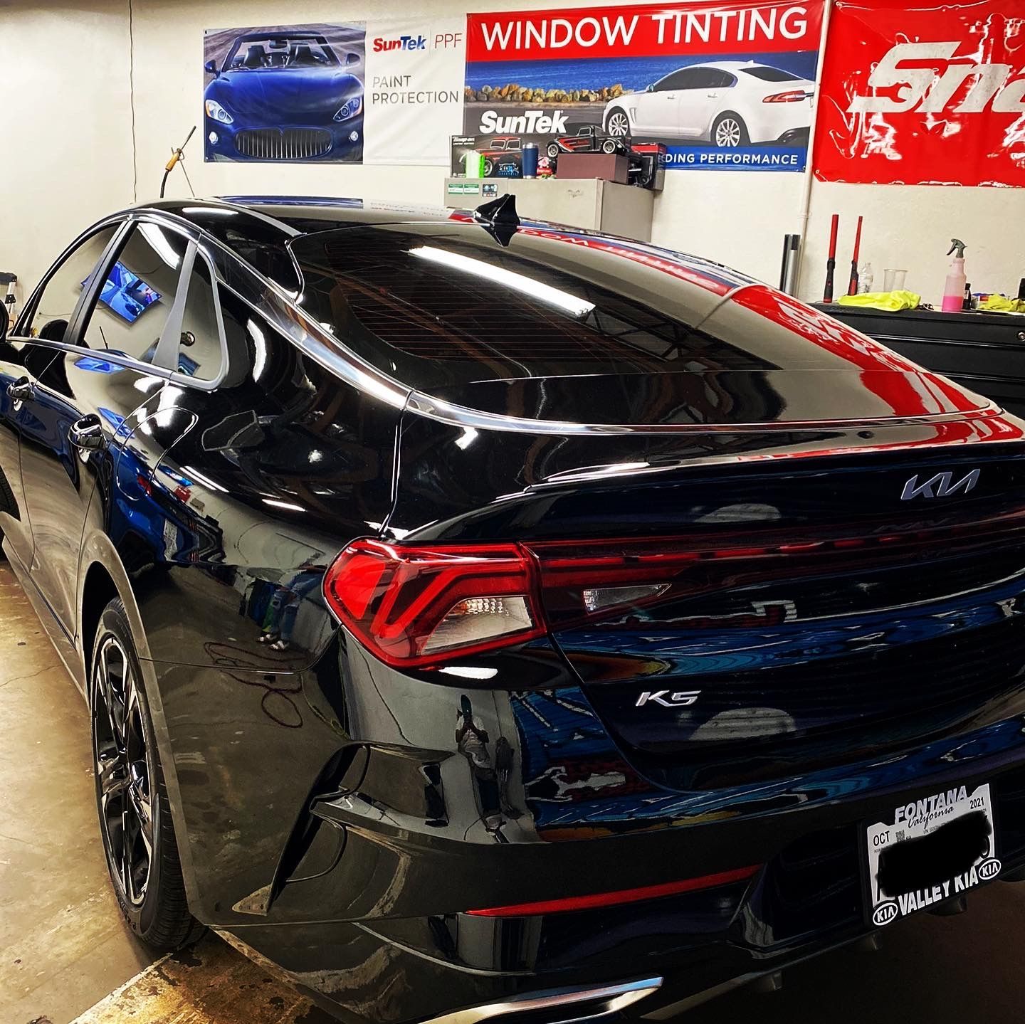 Black Kia K5 sedan in a window tint shop, rear view with glossy paint and red taillights
