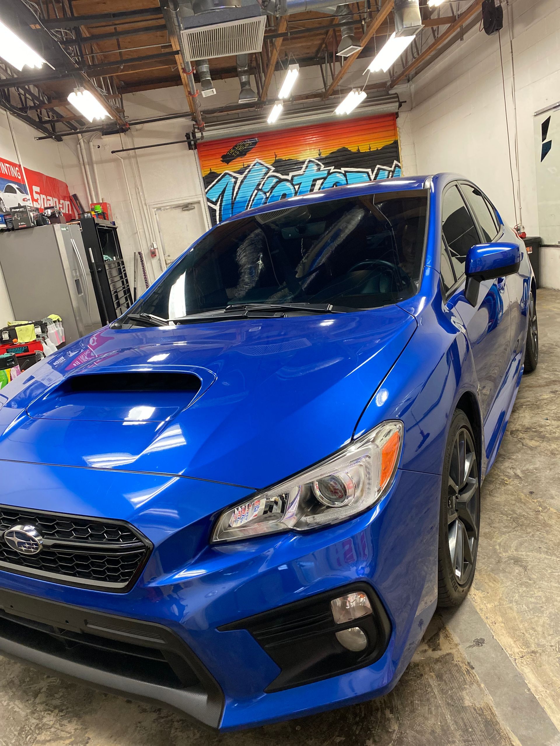 Blue Subaru WRX in an auto shop, seen from the front with graffiti wall in the background.