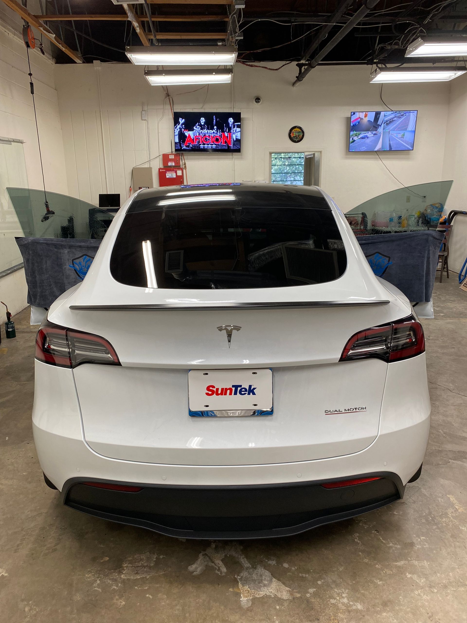 White Tesla in a car wash bay with soap-covered windows and two wall-mounted TVs above.
