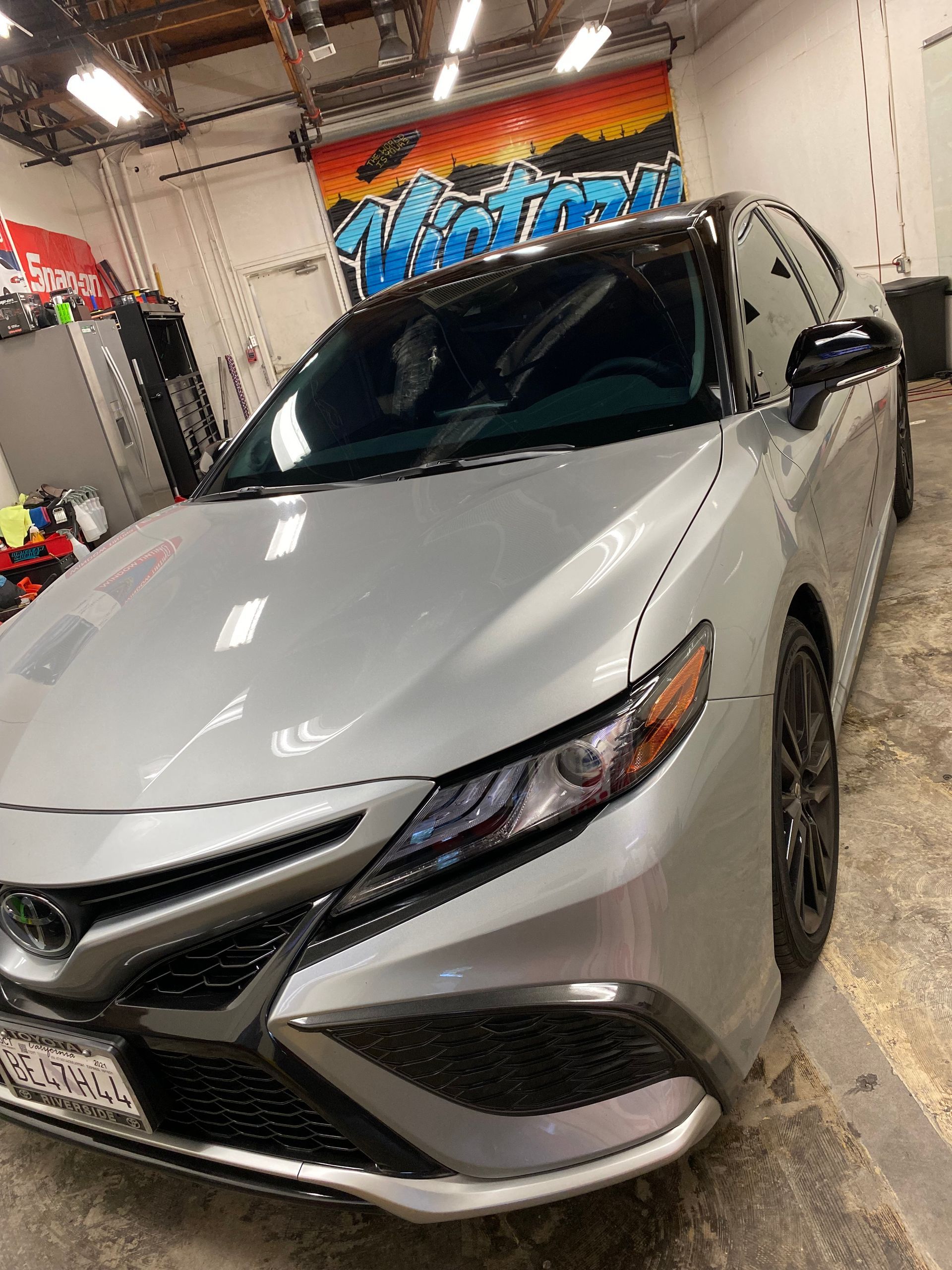 Silver sedan in an auto detailing shop, angled front view with hood and windshield reflecting lights