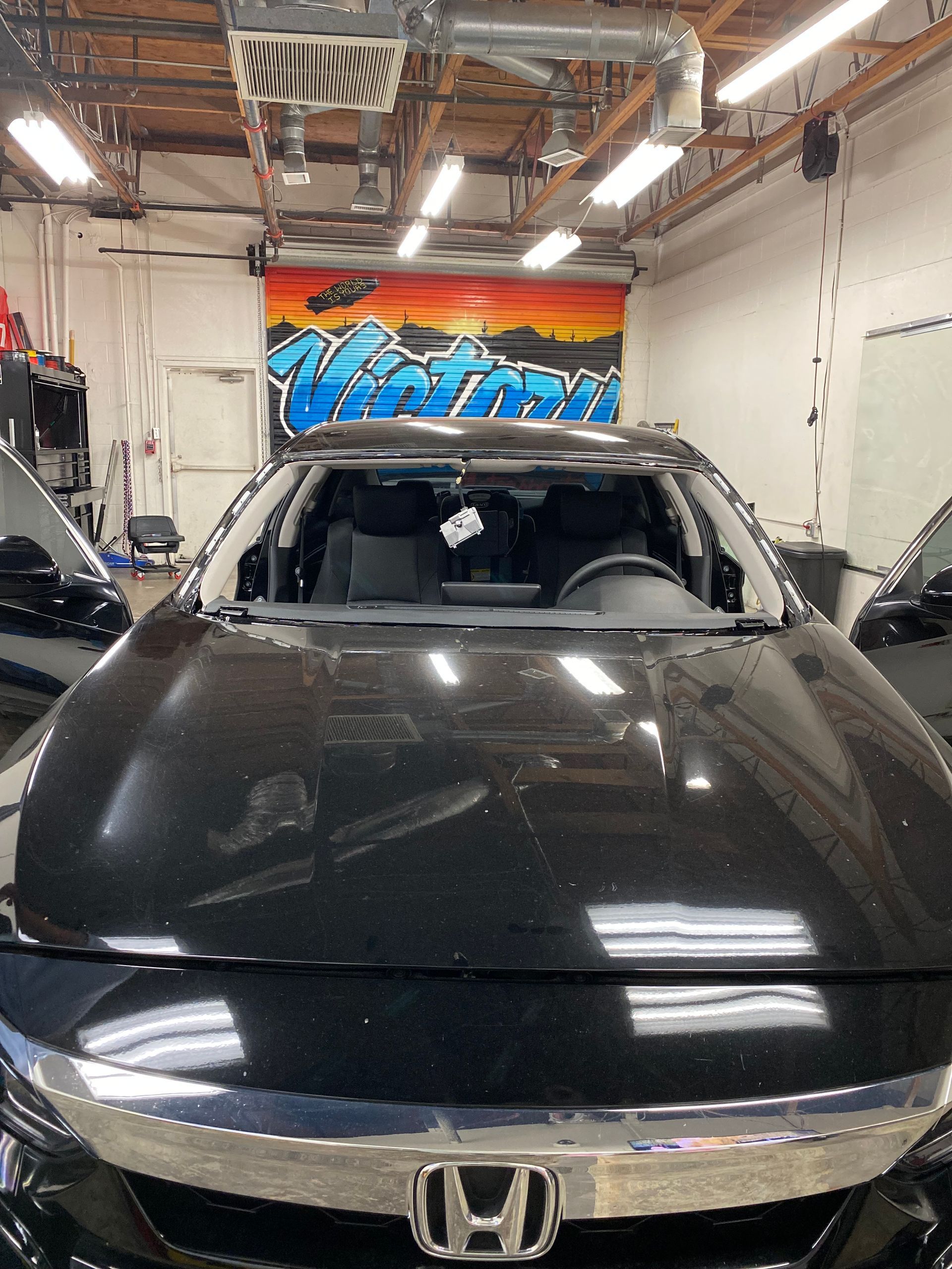 Black Honda sedan in an auto shop, front view with hood open and a colorful wall sign behind it
