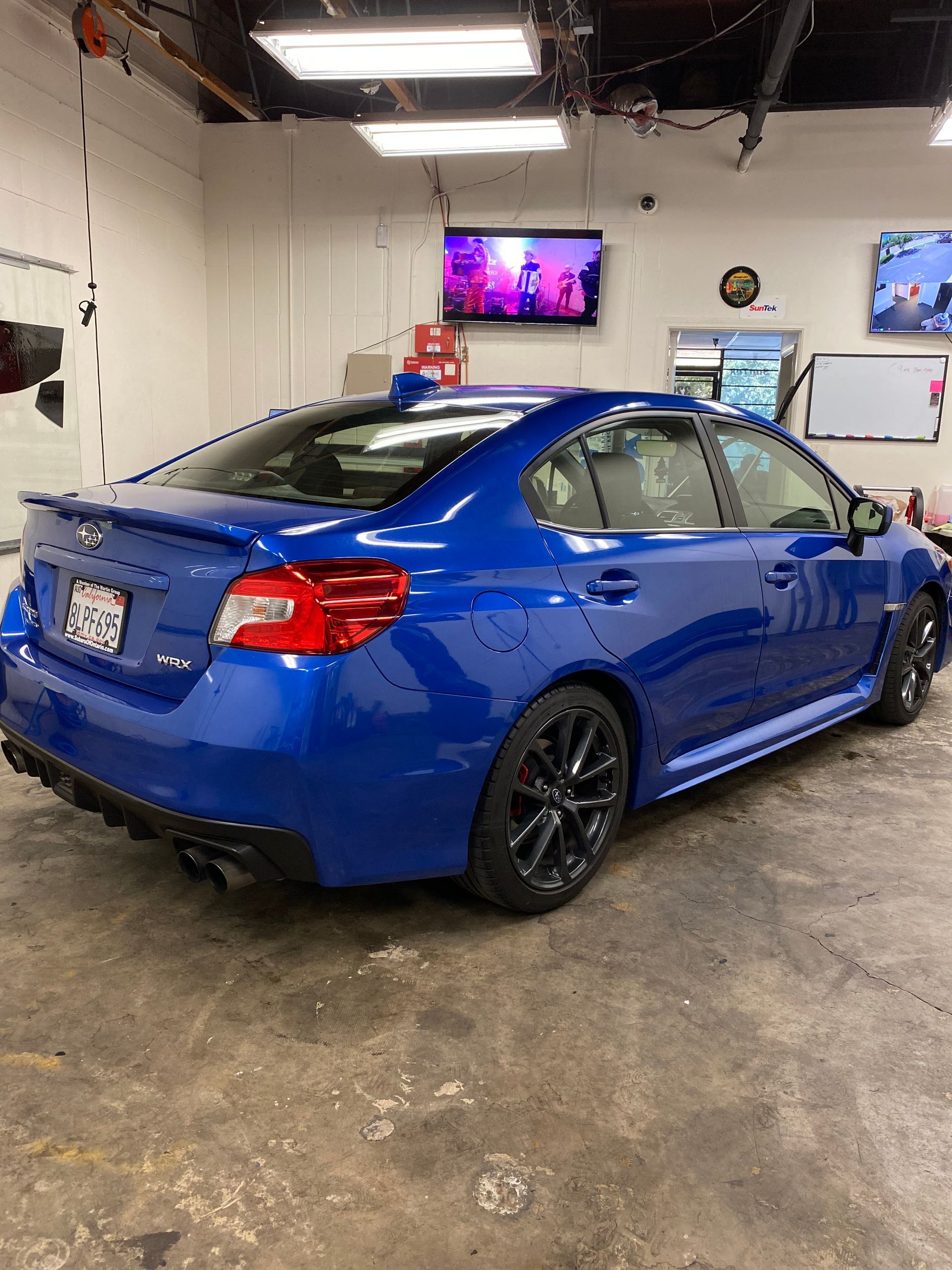 Blue sports sedan in a garage, viewed from the rear-left, with a TV and tools in the background.