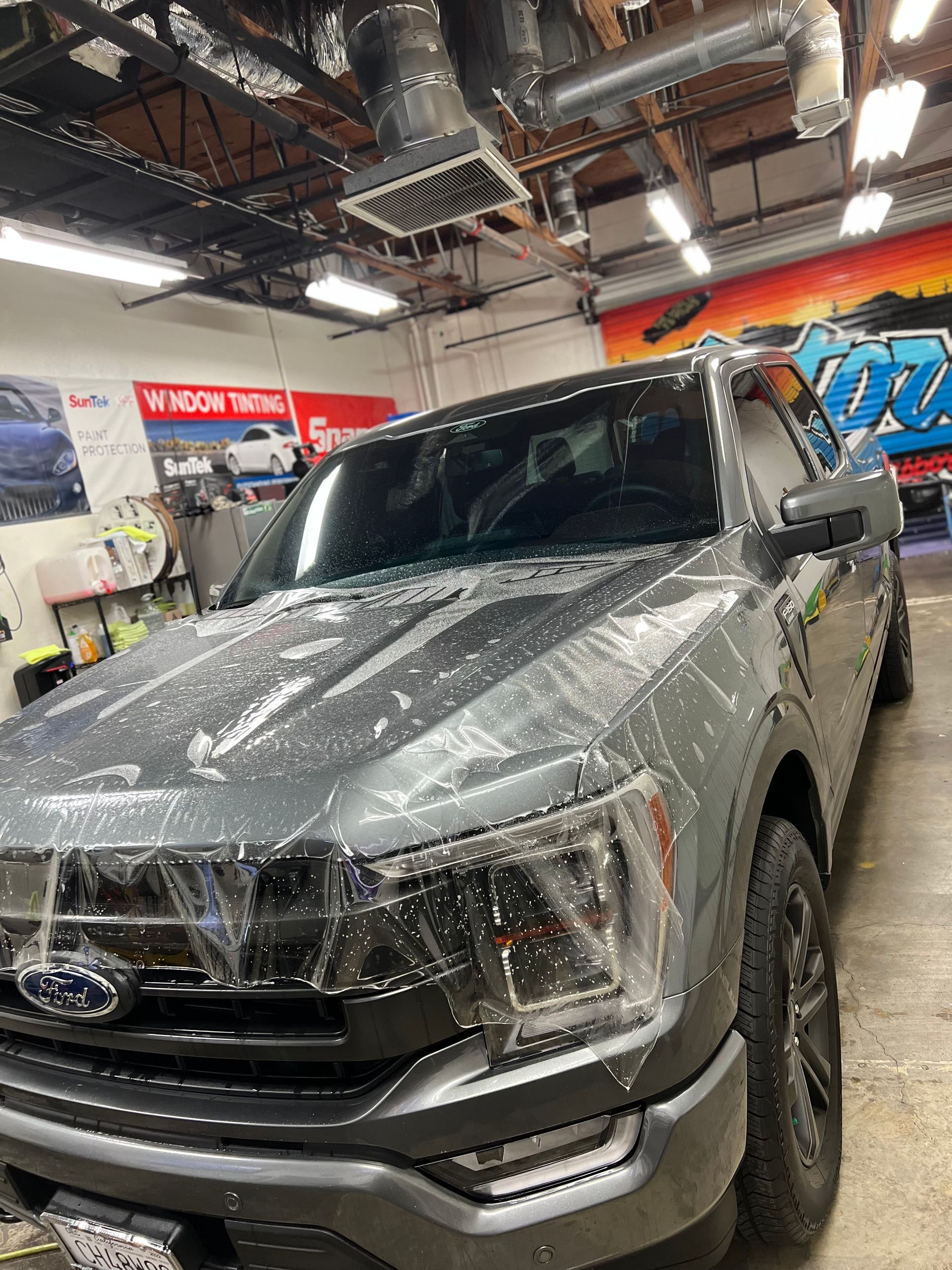 Gray Ford SUV being wrapped in clear film inside an auto shop, with overhead lights and banners visible.