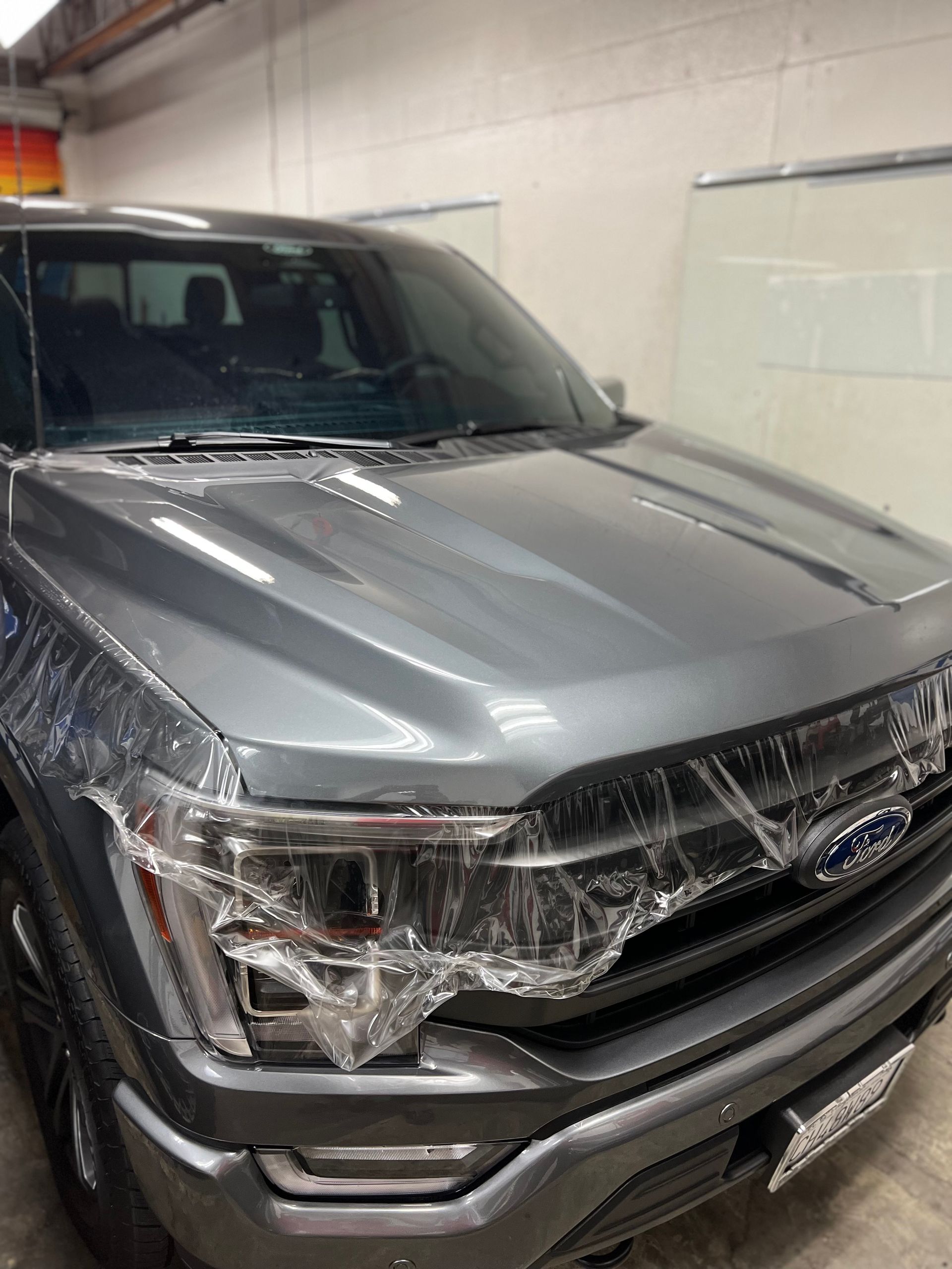 Gray Ford pickup truck with a cracked front headlight and wrapped hood in a garage