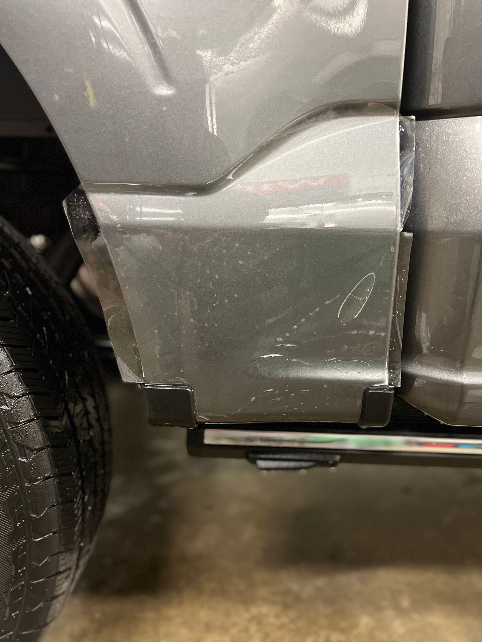 Close-up of a silver car’s rear side panel and wheel near a curb.