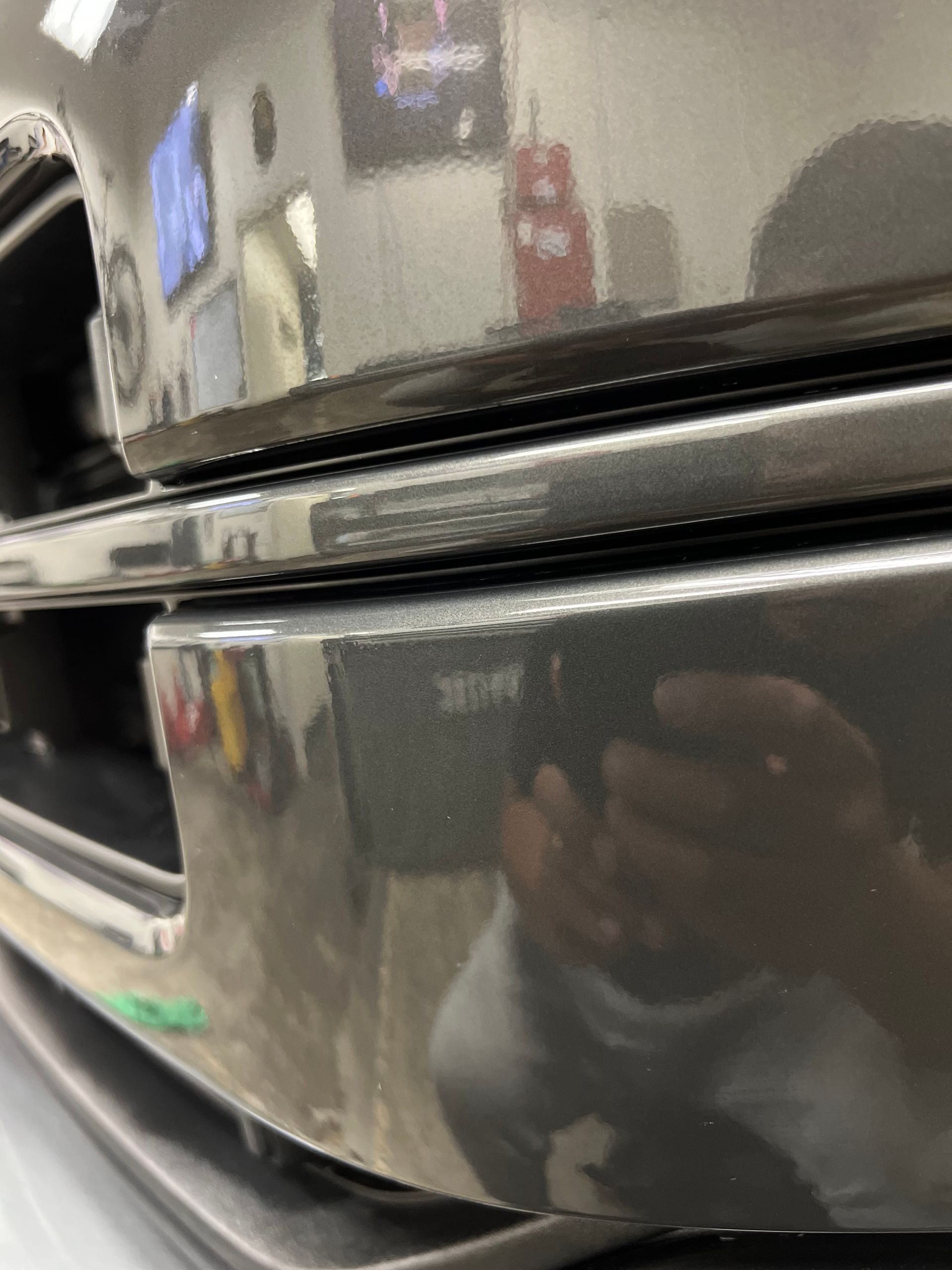Close-up of a glossy black car bumper with a visible reflection of a person holding a phone.