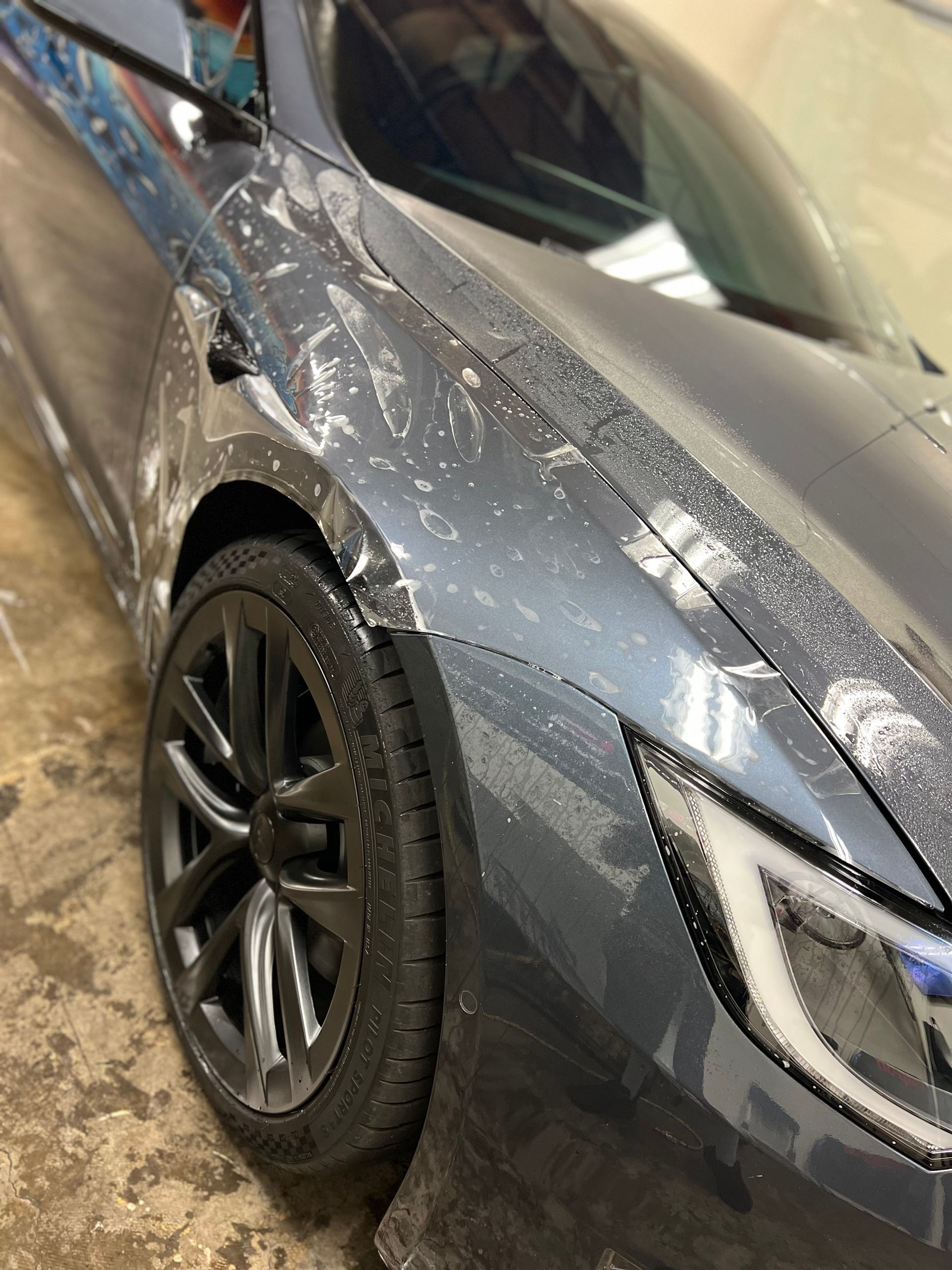 Close-up of a gray car’s front side covered in water droplets during a wash.