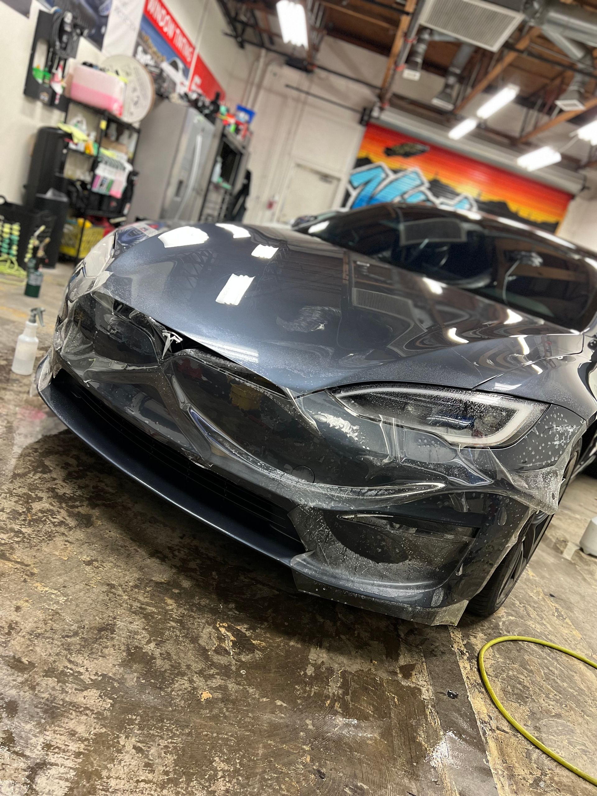 Damaged black Tesla sedan in an auto shop with a crumpled front bumper and hood, wet floor visible.