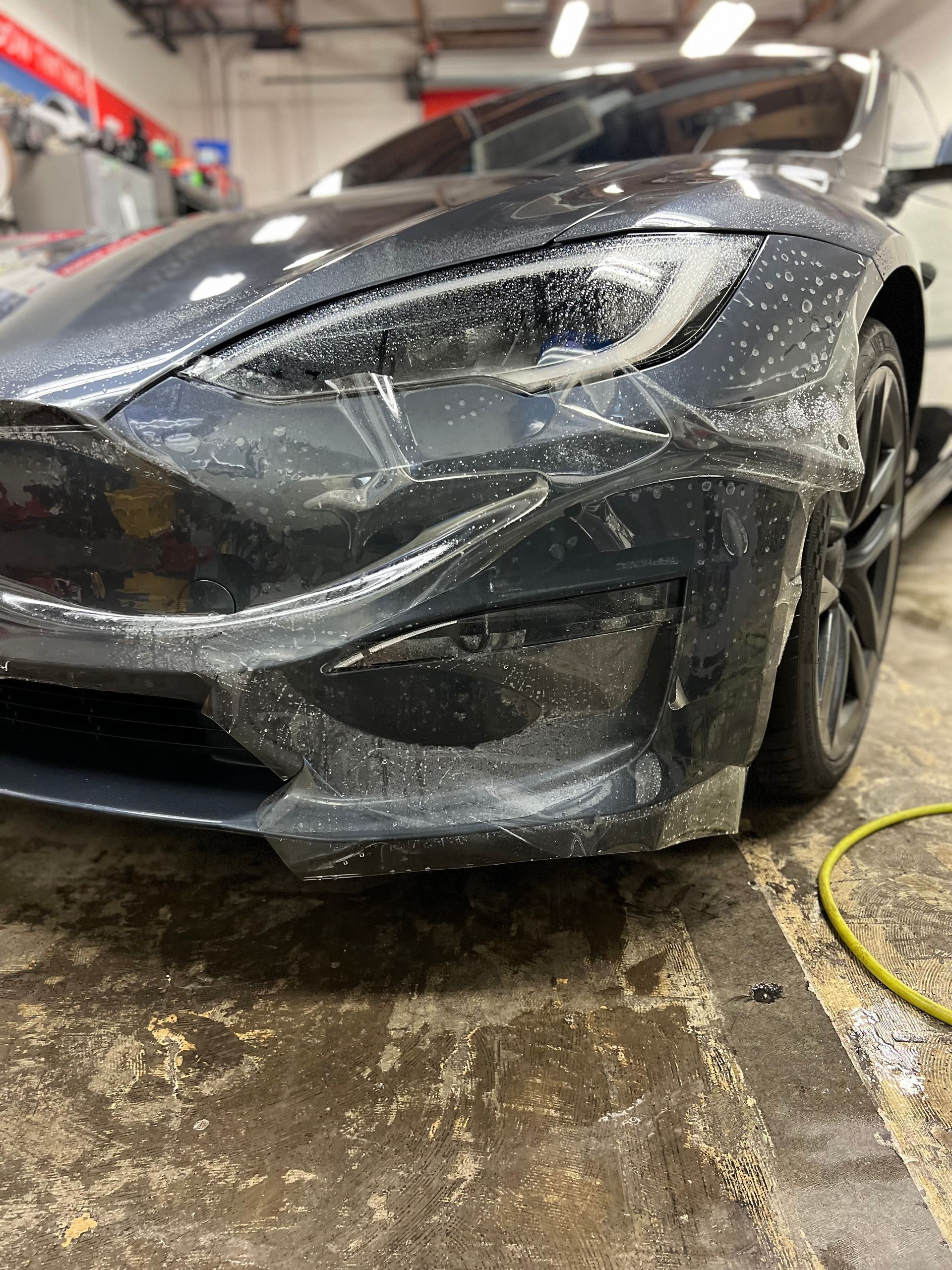 Damaged black sports car in a garage, front bumper and hood covered in plastic wrap