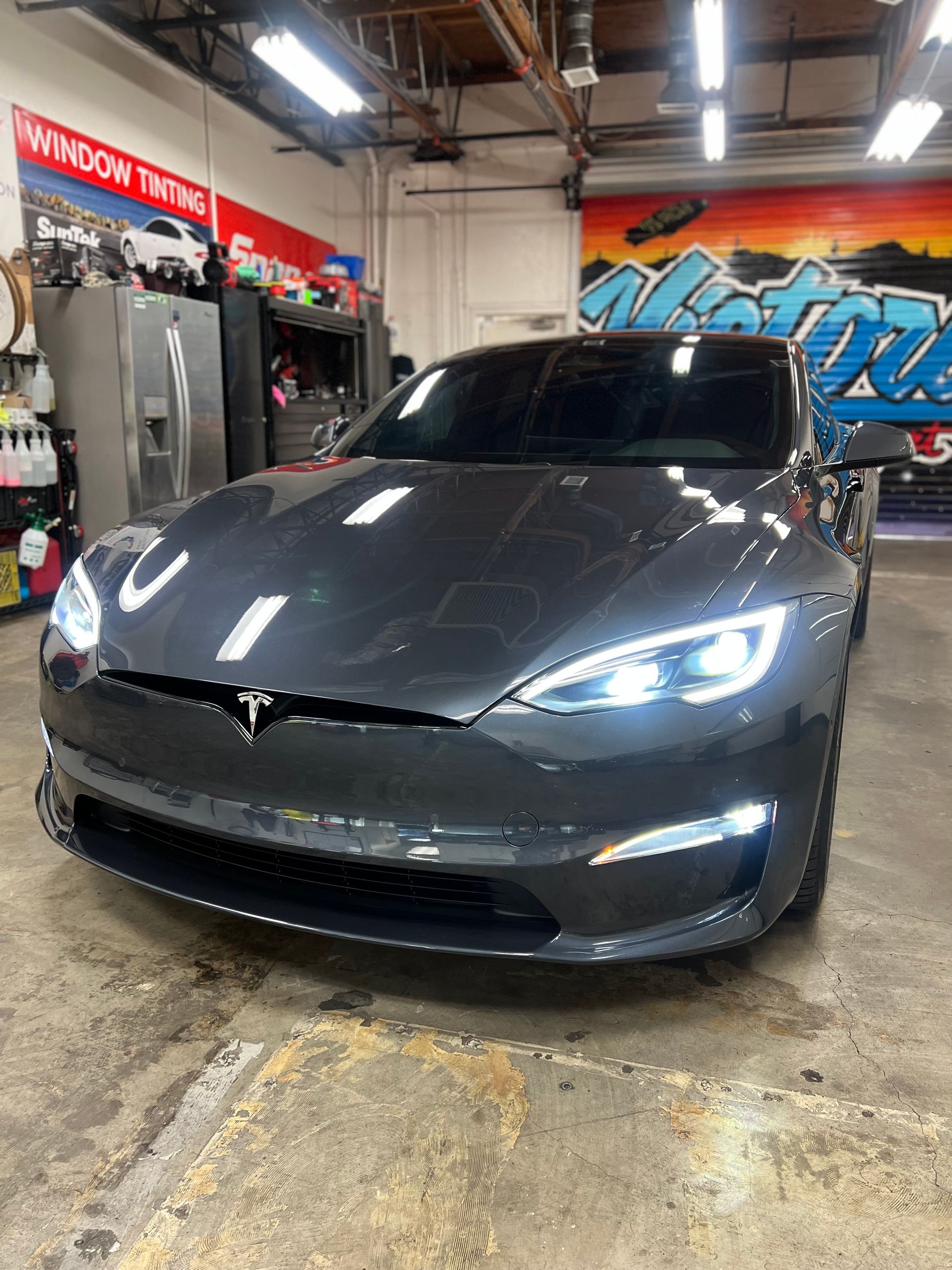 Gray Tesla sedan in an auto shop, viewed from the front with headlights on.