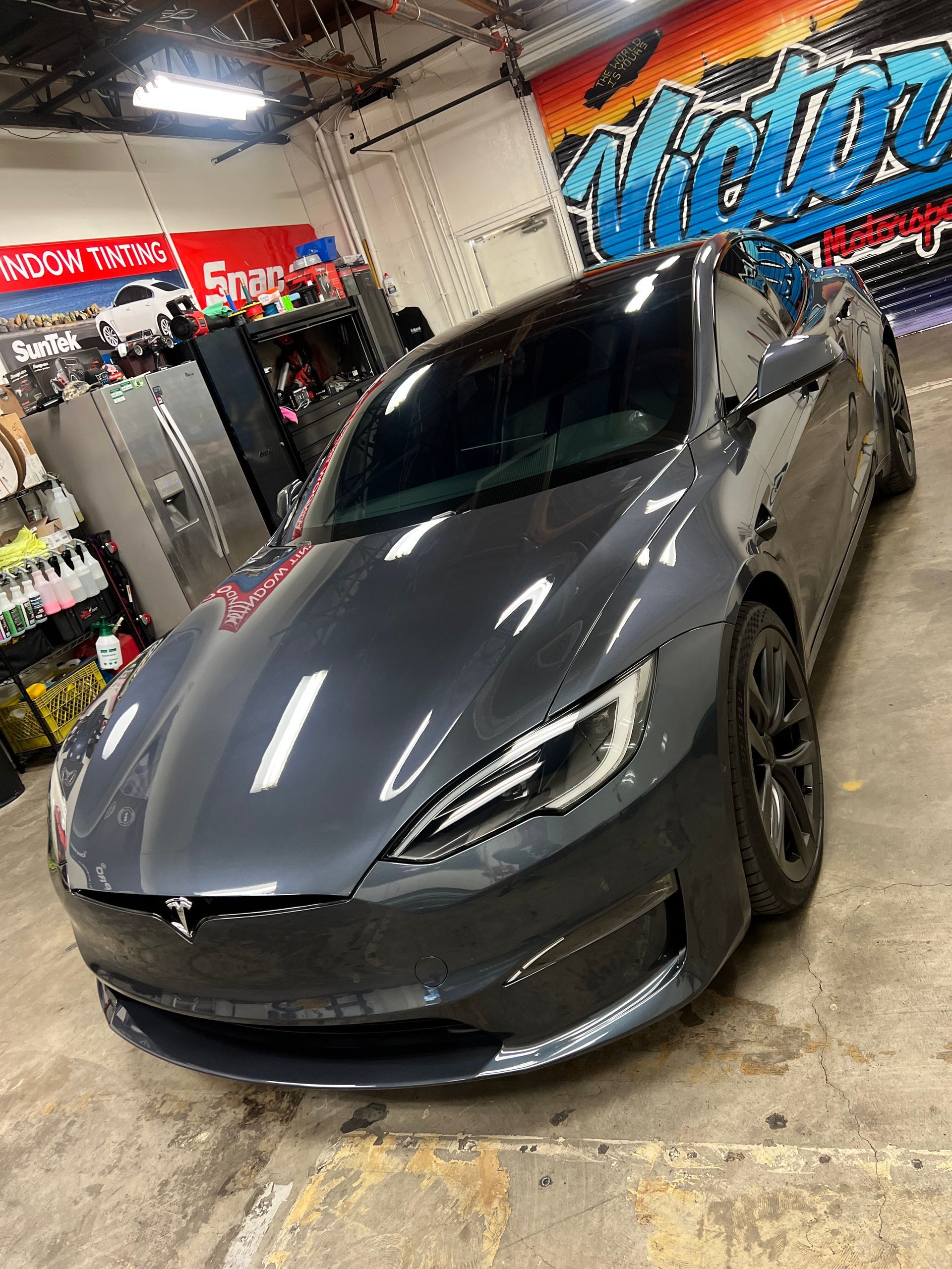 Gray sports car parked inside an auto shop with bright overhead lights and tire displays in the background