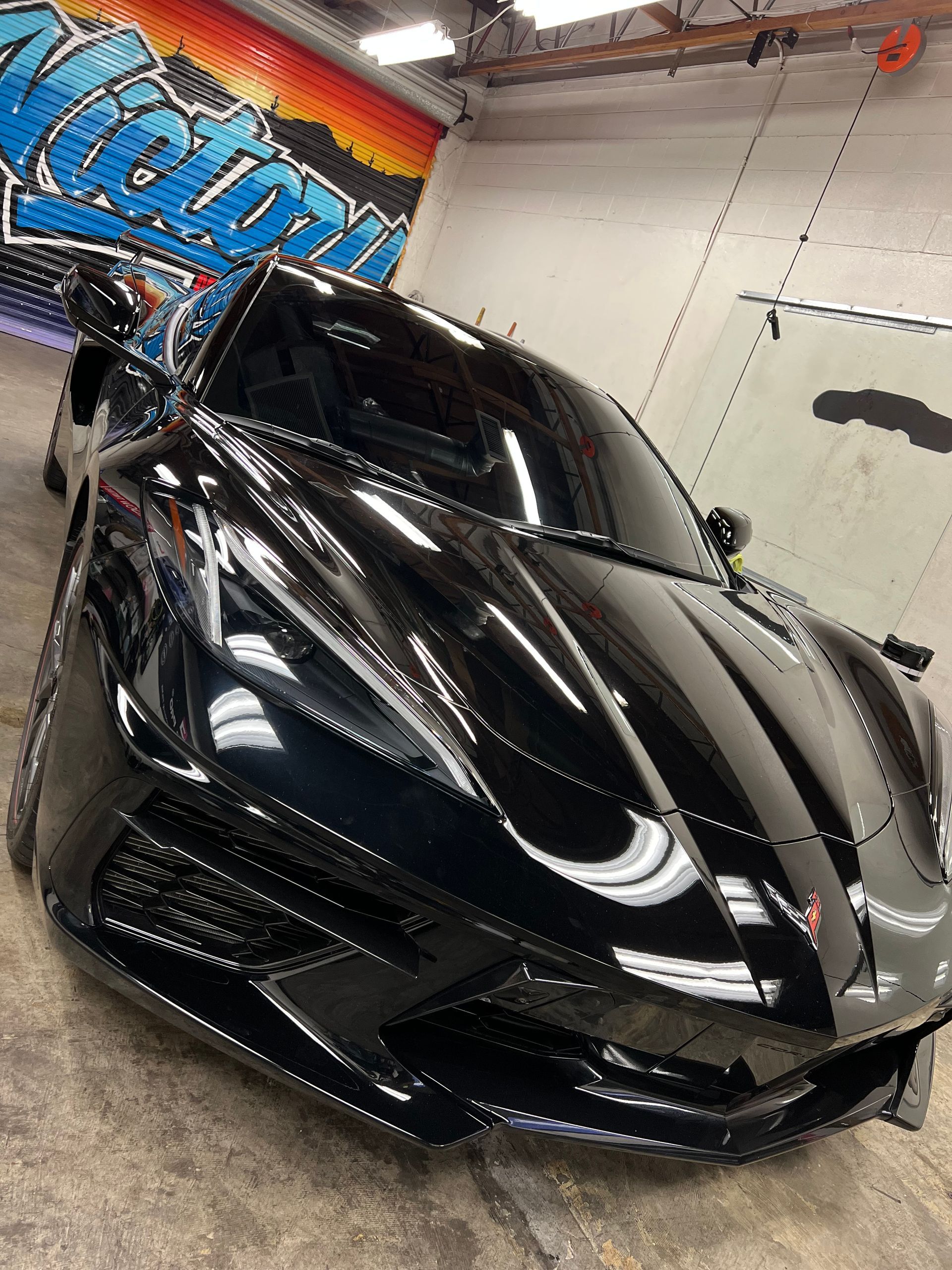 Black sports car with glossy stripes in a garage, angled front view with headlights on